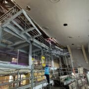 A construction worker stands on scaffolding inside a large building under renovation, with exposed pipes and partially finished walls visible.