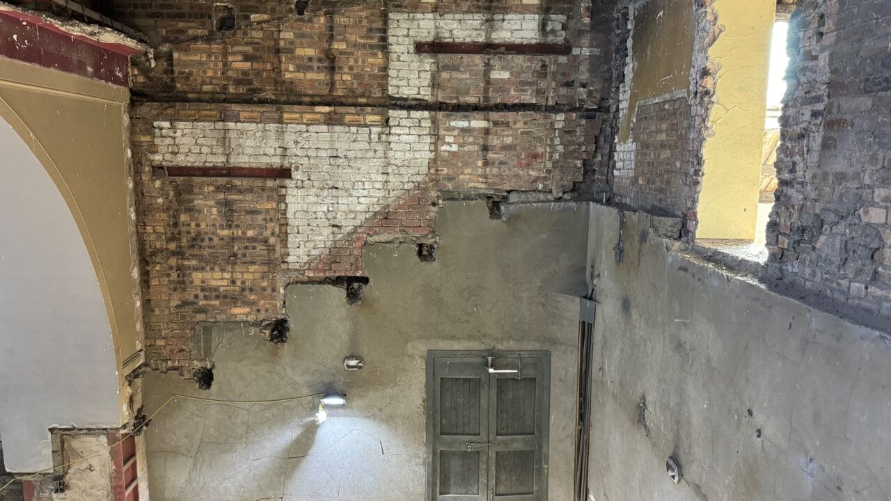 Interior view of a partially renovated brick wall with exposed areas, an old door, a large open window, and visible construction materials.
