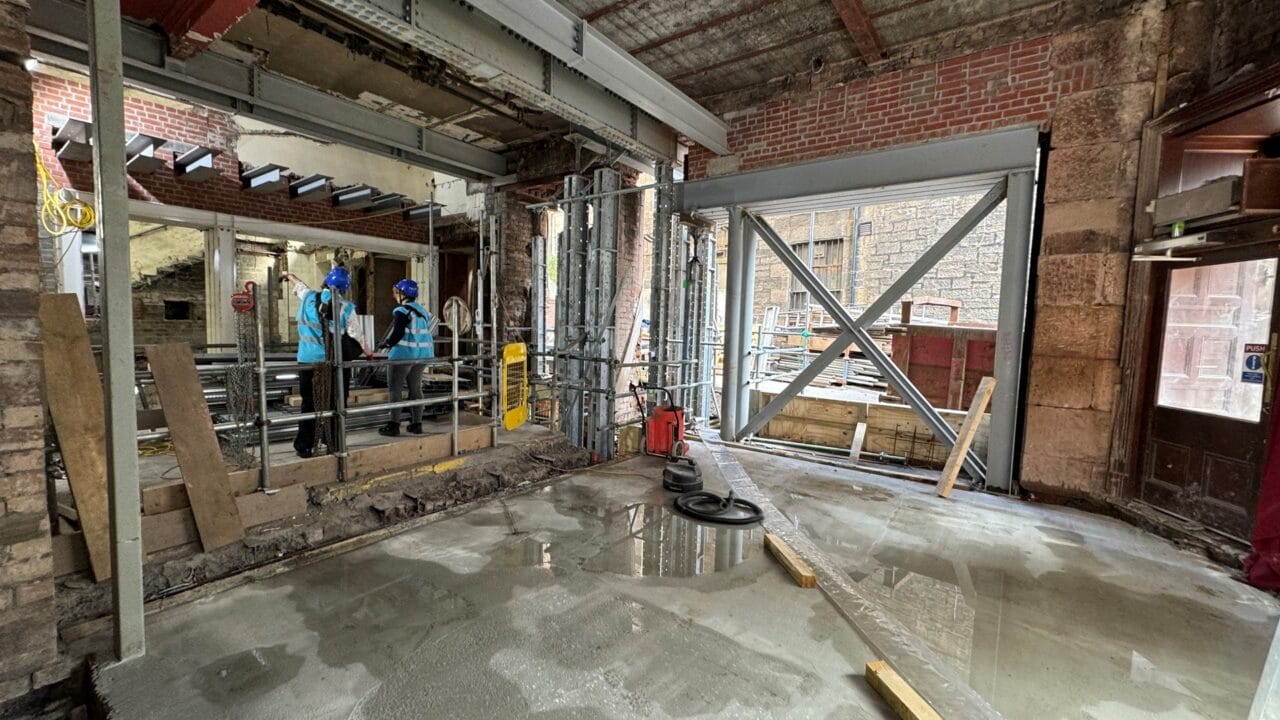 Construction site interior with exposed brick walls, metal beams, construction materials, and two workers in blue safety gear standing by a railing.