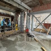 Construction site interior with exposed brick walls, metal beams, construction materials, and two workers in blue safety gear standing by a railing.