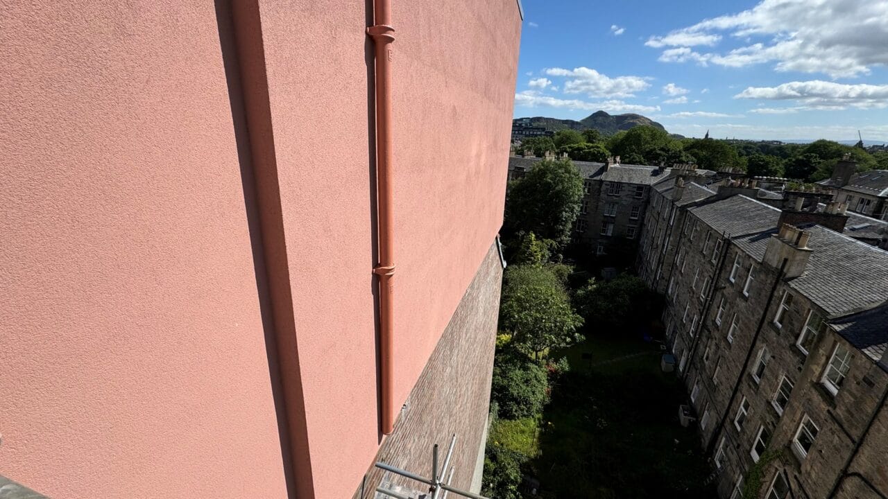 View of a pink building wall overlooking a courtyard with grass, trees, and adjacent stone buildings, under a blue sky with scattered clouds. Hills are visible in the distance.