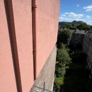 View of a pink building wall overlooking a courtyard with grass, trees, and adjacent stone buildings, under a blue sky with scattered clouds. Hills are visible in the distance.