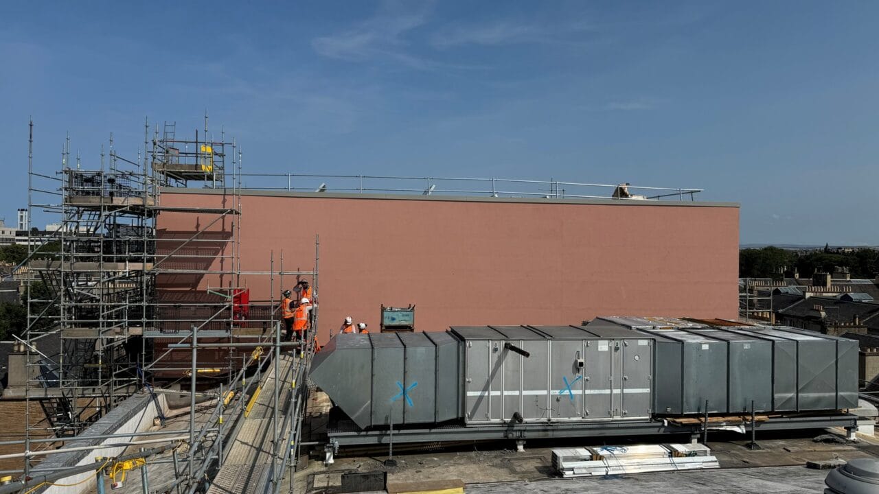 Construction workers in orange vests stand near scaffolding on a rooftop next to large HVAC units, with a tall pink wall and clear sky in the background.