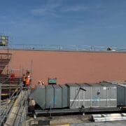 Construction workers in orange vests stand near scaffolding on a rooftop next to large HVAC units, with a tall pink wall and clear sky in the background.