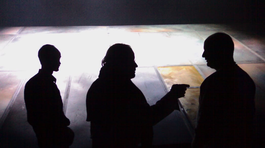 Three people are seen in silhouette, standing indoors on a tiled floor, with one person gesturing toward another.