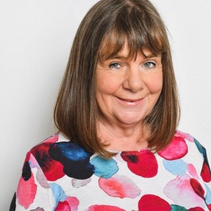 A middle-aged woman with straight brown hair smiles at the camera, wearing a white shirt with a colorful polka dot pattern, as if in the spotlight against a plain white background.