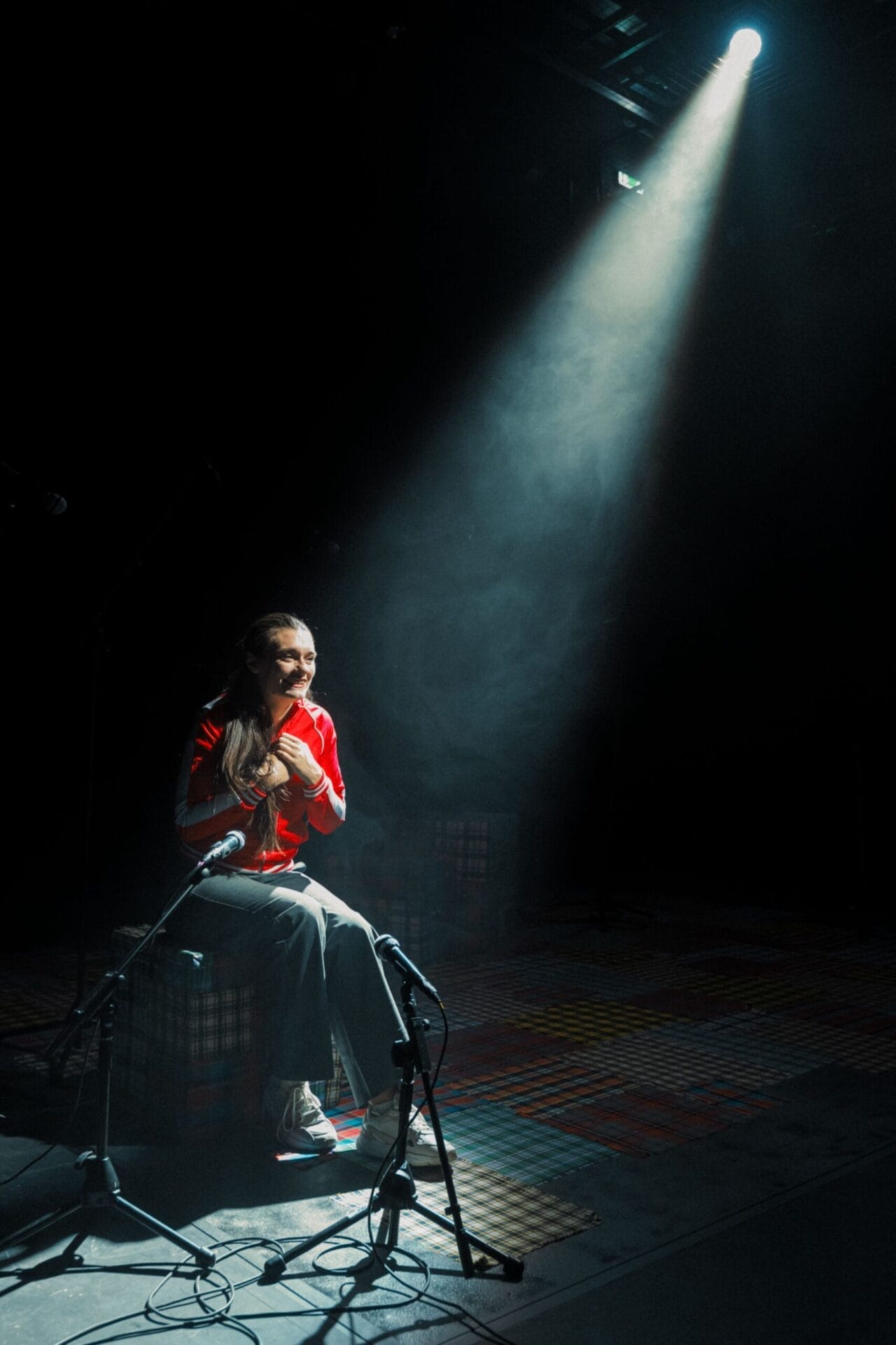 A person in a red shirt sits on a crate under a bright spotlight on a dimly lit stage, poised to speak into two microphones—ready to share stories in the Common Tongue.