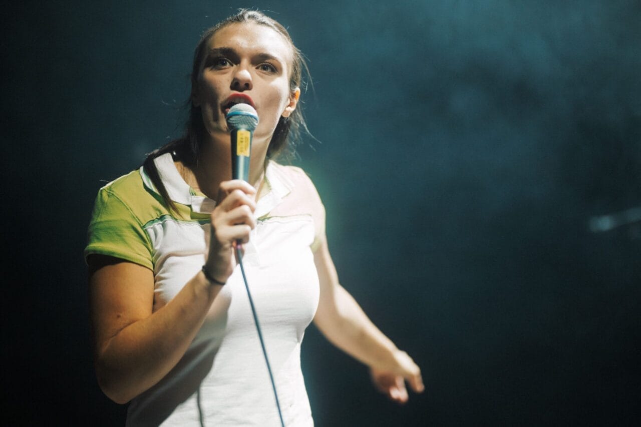 A woman holding a microphone stands on stage under dramatic lighting, looking forward and pointing with her left hand as if inviting the audience to join her in the Common Tongue.