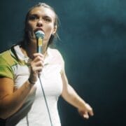 A woman holding a microphone stands on stage under dramatic lighting, looking forward and pointing with her left hand as if inviting the audience to join her in the Common Tongue.