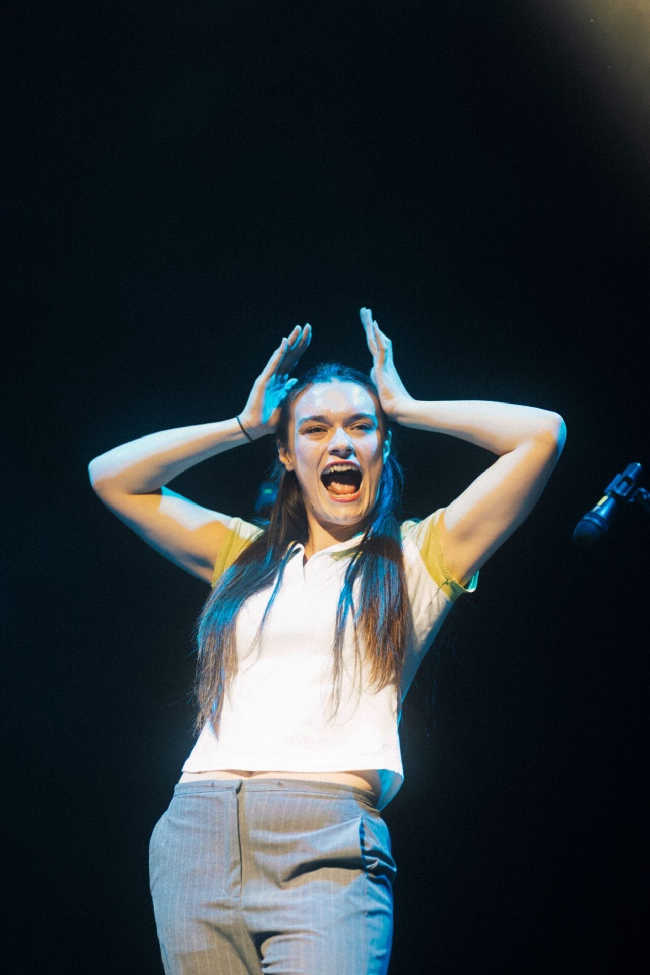 A person with long hair stands on stage under a spotlight, holding their hands near their head and expressing excitement or surprise. A microphone, ready for the Common Tongue performance, is visible at the right edge.
