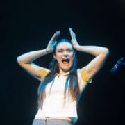 A person with long hair stands on stage under a spotlight, holding their hands near their head and expressing excitement or surprise. A microphone, ready for the Common Tongue performance, is visible at the right edge.