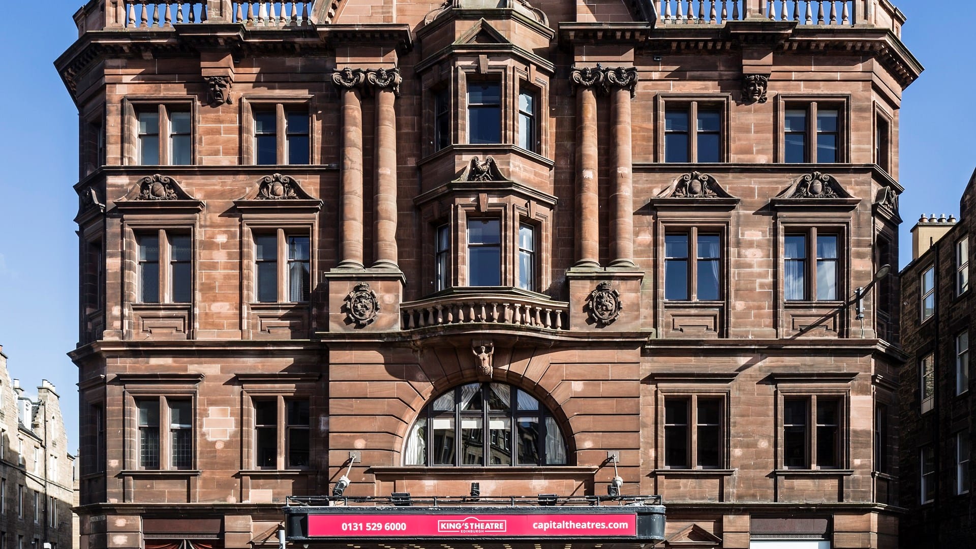 A symmetrical, historic stone theatre building with ornate columns, arched windows, and a red sign reading “King’s Theatre” above the entrance.