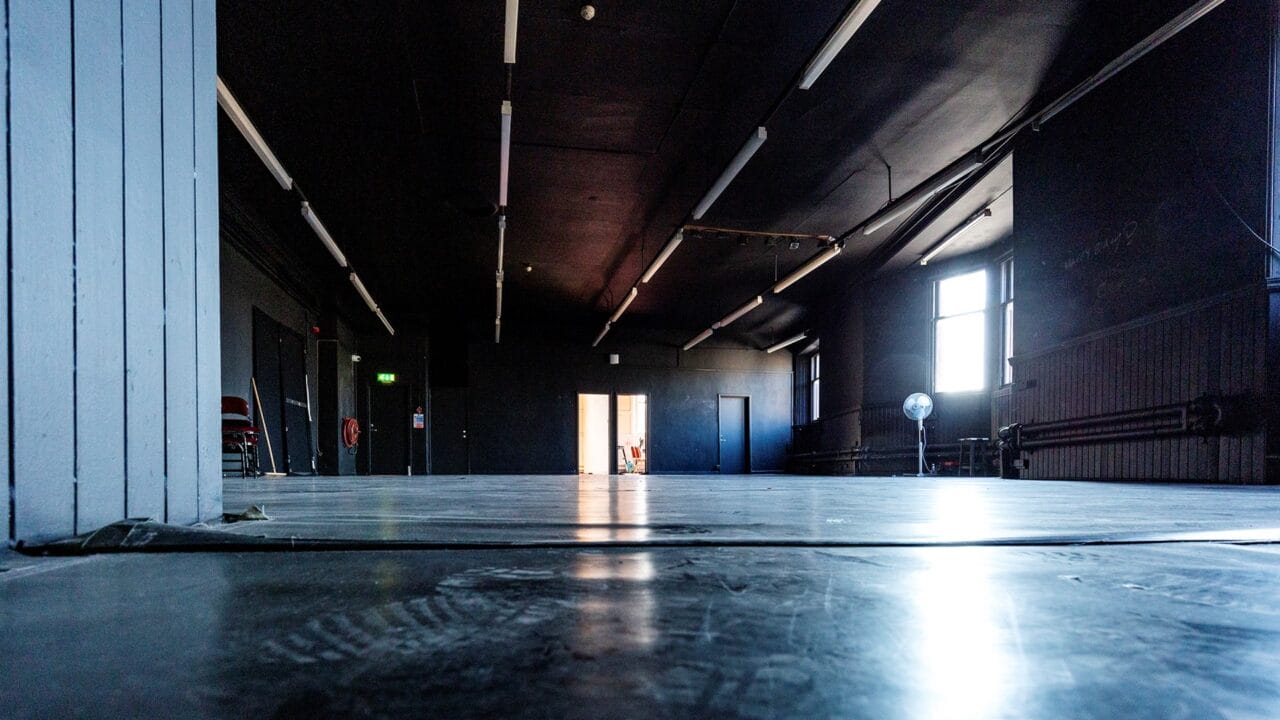 Low-angle view of a large, empty room with dark walls and ceiling, fluorescent lights, a fan, a few stacked chairs, and light coming through an open door at the far end.