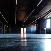 Low-angle view of a large, empty room with dark walls and ceiling, fluorescent lights, a fan, a few stacked chairs, and light coming through an open door at the far end.