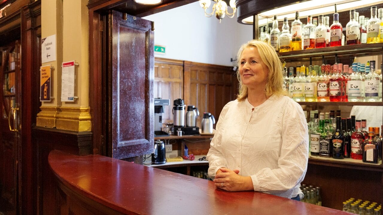 A woman in a white blouse stands behind a wooden bar counter, with shelves of liquor bottles and coffee machines visible in the background.