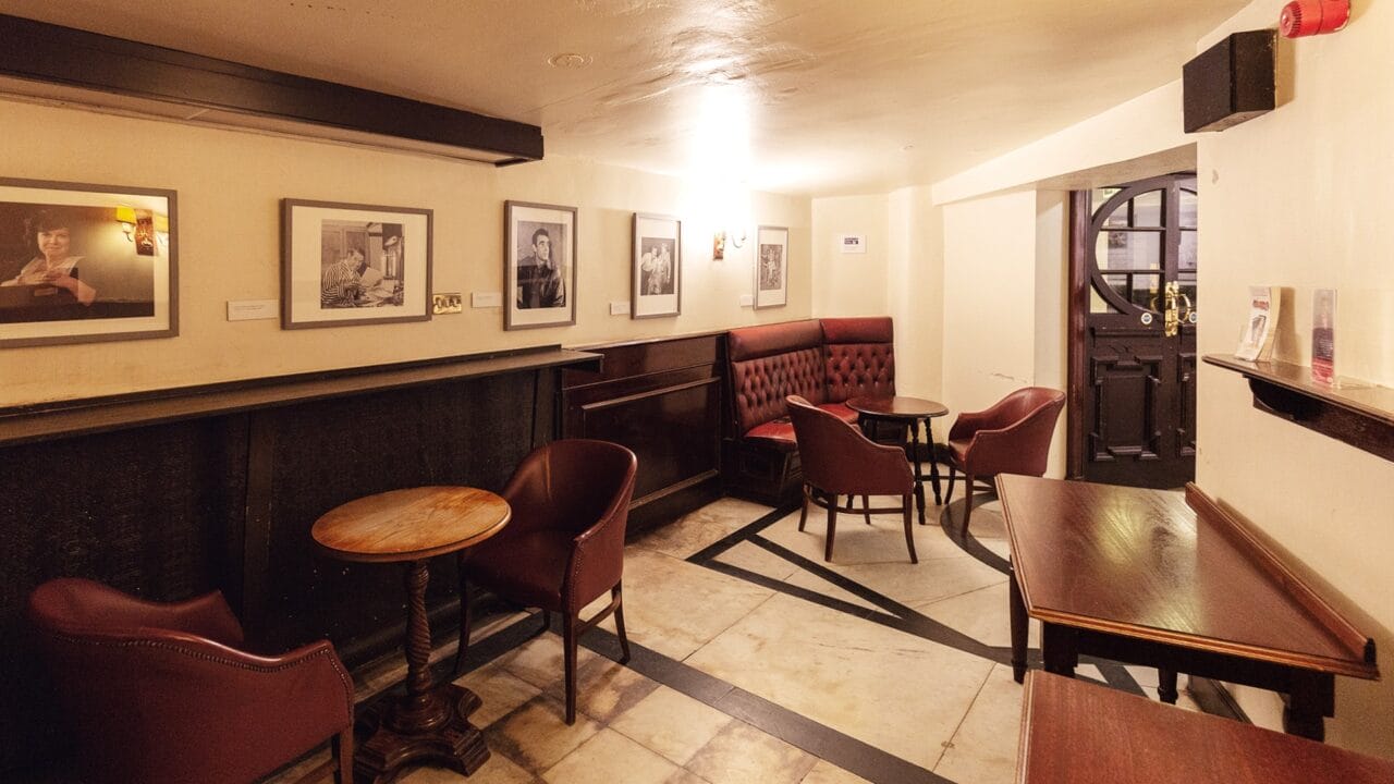 Small, dimly lit café or pub with framed photos on cream walls, round wooden tables, and cushioned chairs arranged around the room.