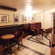 Small, dimly lit café or pub with framed photos on cream walls, round wooden tables, and cushioned chairs arranged around the room.