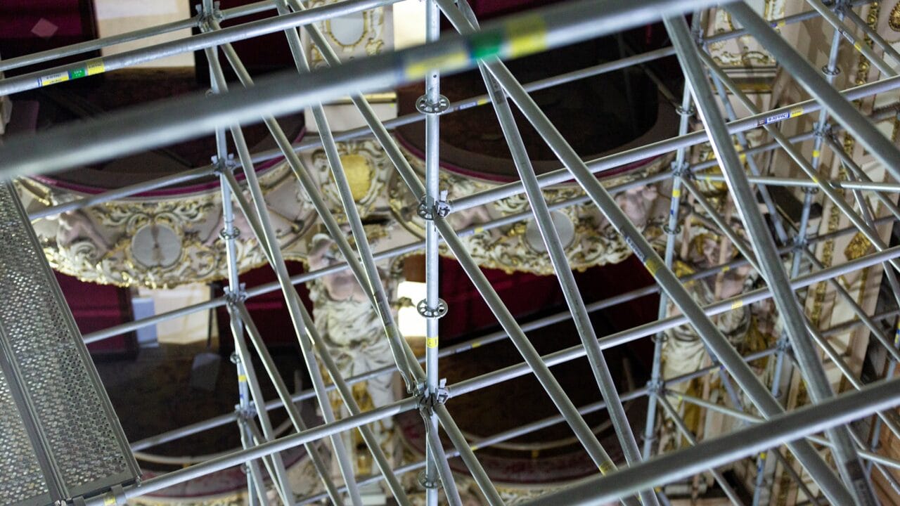 Metal scaffolding bars intersect in front of an ornate balcony with decorative moldings and red velvet seats, indicating ongoing restoration or maintenance work.