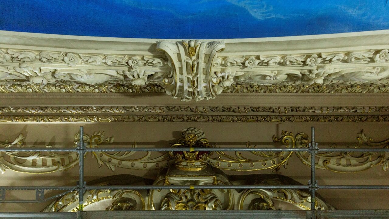 Ornate architectural ceiling with intricate moldings and gold accents, partially obscured by metal scaffolding.