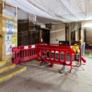Construction site interior with wooden stairs, safety signage, red barriers blocking access, and plastic sheeting covering the ceiling.