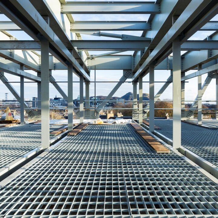 Steel framework and grated flooring of a building under construction, with an urban landscape visible in the background.