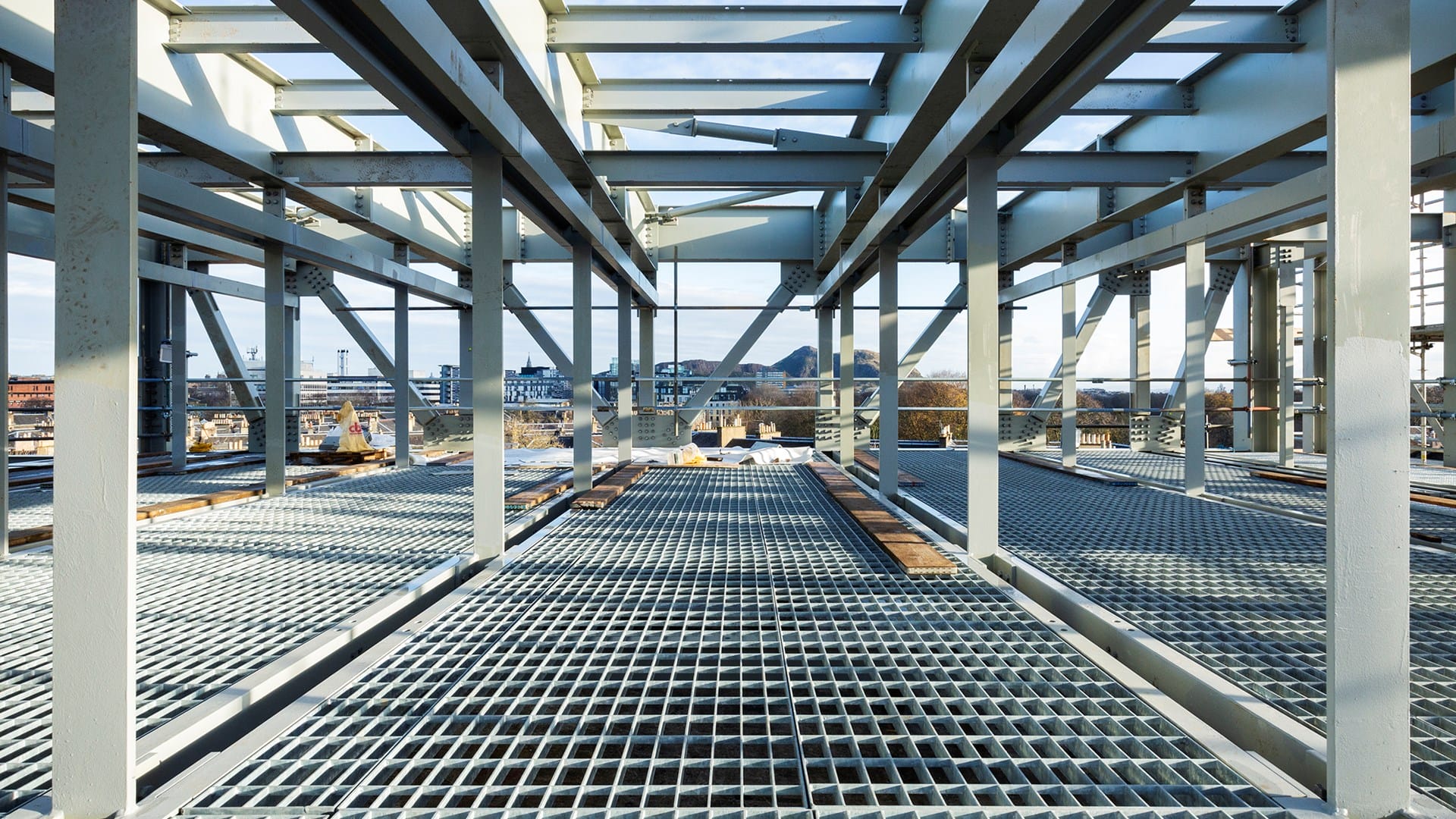 Steel framework and grated flooring of a building under construction, with an urban landscape visible in the background.