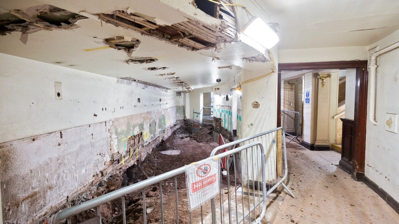 An interior hallway under construction with exposed dirt, damaged walls and ceiling, safety barriers, and construction signs.