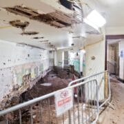 An interior hallway under construction with exposed dirt, damaged walls and ceiling, safety barriers, and construction signs.