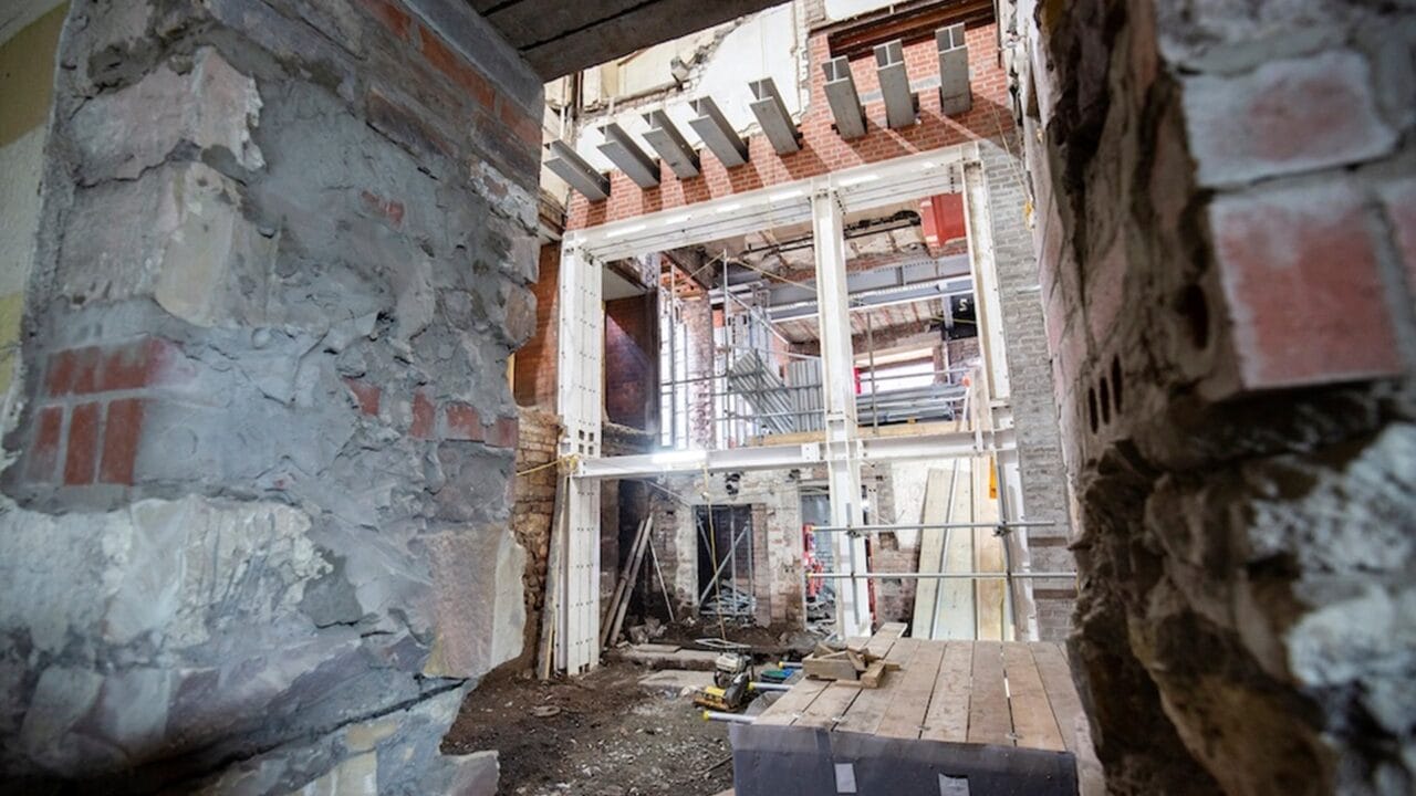 Partially demolished interior of a building under renovation, with exposed brick walls, steel beams, scaffolding, and construction materials scattered on the ground.