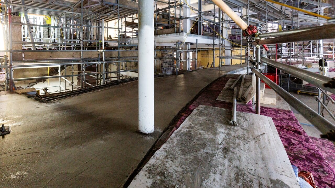 A construction site with scaffolding and freshly poured concrete on a curved ramp or floor; a white structural column stands in the middle.