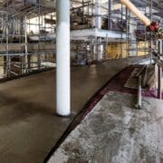 A construction site with scaffolding and freshly poured concrete on a curved ramp or floor; a white structural column stands in the middle.