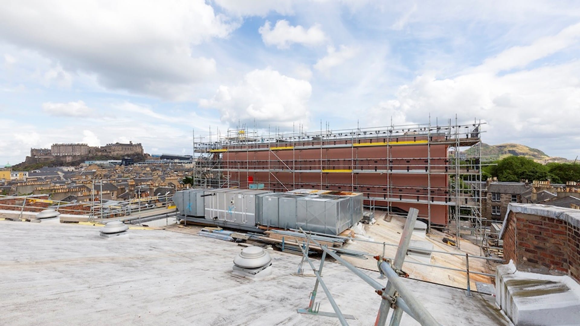 A building under construction with scaffolding, seen from a rooftop, with a cityscape and historic castle in the background under a cloudy sky.
