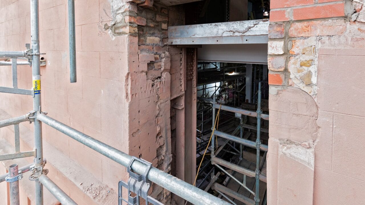 A close-up view of a partially demolished brick and stone wall, showing exposed scaffolding and interior construction materials.