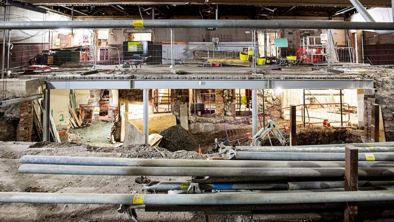 Construction site with exposed foundation, steel beams, scaffolding, dirt, and building materials; partially demolished interior visible in the background.