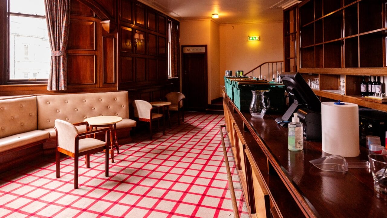 A quiet bar interior with wood paneling, upholstered seating, a checkered carpet, and an empty counter with various utensils and supplies.