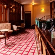 A quiet bar interior with wood paneling, upholstered seating, a checkered carpet, and an empty counter with various utensils and supplies.