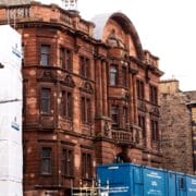 A red sandstone building with ornate architectural details stands between stone buildings, with blue construction containers and scaffolding in front.