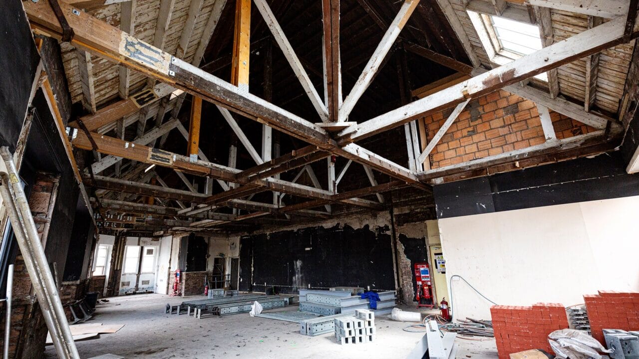 Interior of a building under renovation with exposed beams, unfinished walls, metal and brick materials, and construction equipment scattered on the floor.