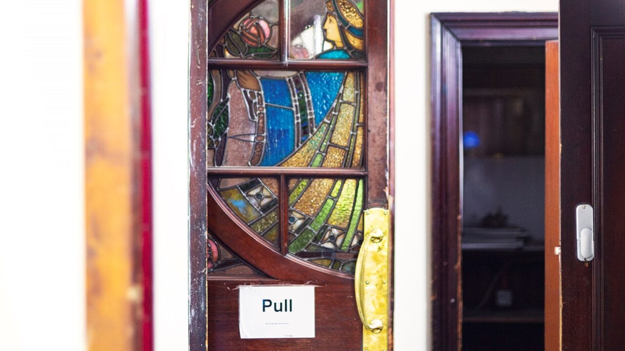 A stained glass door with an image of a person holding a flower has a paper sign reading "Pull" taped to it. The door is partially open, revealing a room beyond.