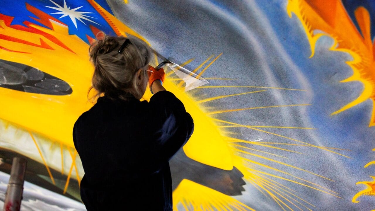 A person in gloves restores a colorful ceiling mural, using a small tool to clean or repair the painted surface.