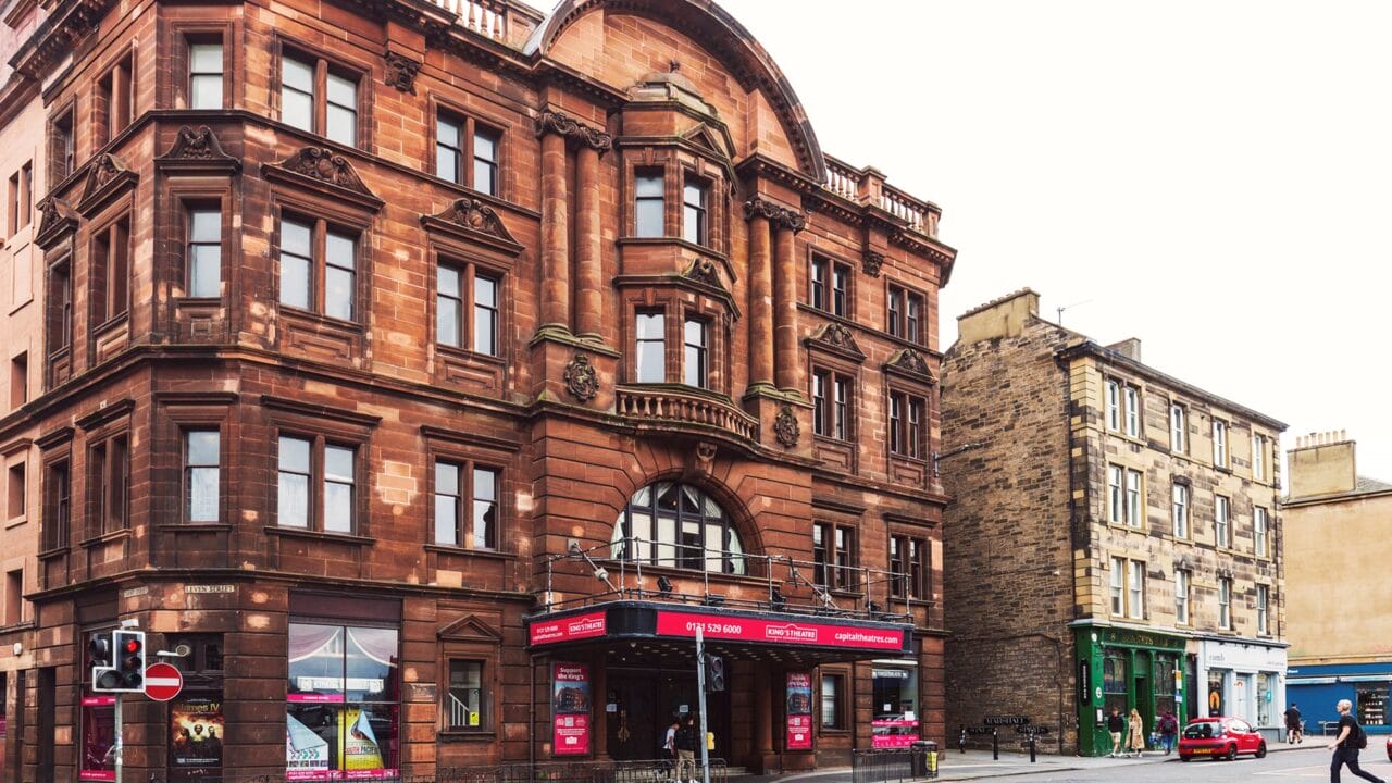 A historic red sandstone building with large arched windows houses a theater; adjacent is a beige brick building with shops at street level. People and cars are visible on the street.