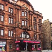 A historic red sandstone building with large arched windows houses a theater; adjacent is a beige brick building with shops at street level. People and cars are visible on the street.