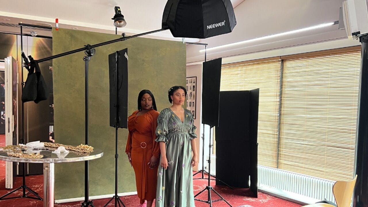 Danielle Jam and another woman stand side by side in a photography studio, under the spotlight, with lighting equipment, a green backdrop, and a table displaying jewelry in the foreground.
