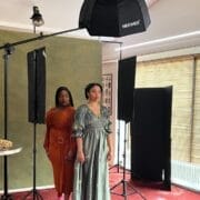 Danielle Jam and another woman stand side by side in a photography studio, under the spotlight, with lighting equipment, a green backdrop, and a table displaying jewelry in the foreground.