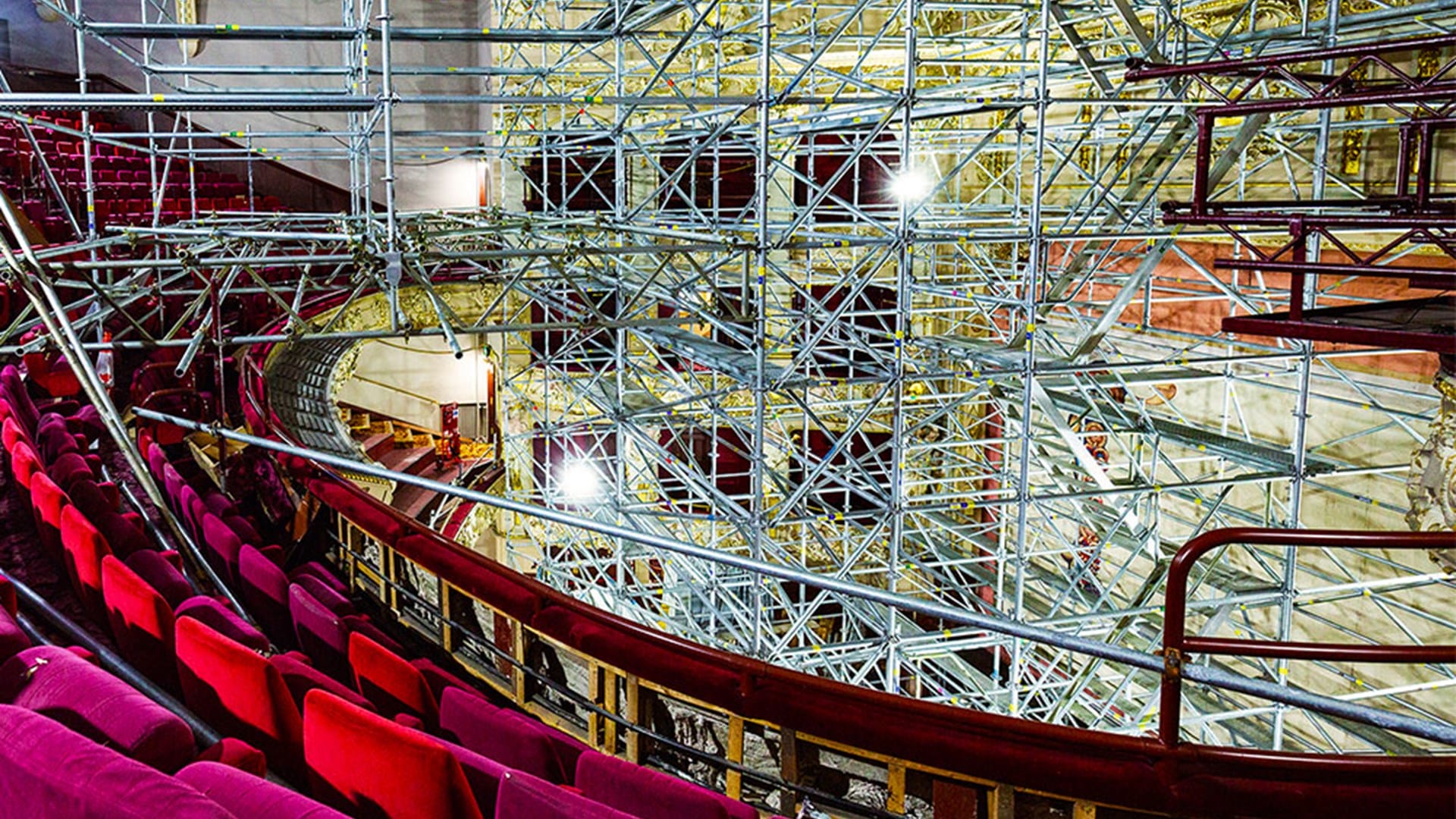 Rows of red theater seats face a stage filled with extensive metal scaffolding inside an ornate, under-renovation theater.