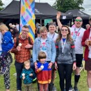 A group of people, some wearing medals and race bibs, pose outdoors at an event with tents and a crowd in the background. Two children stand at the front.