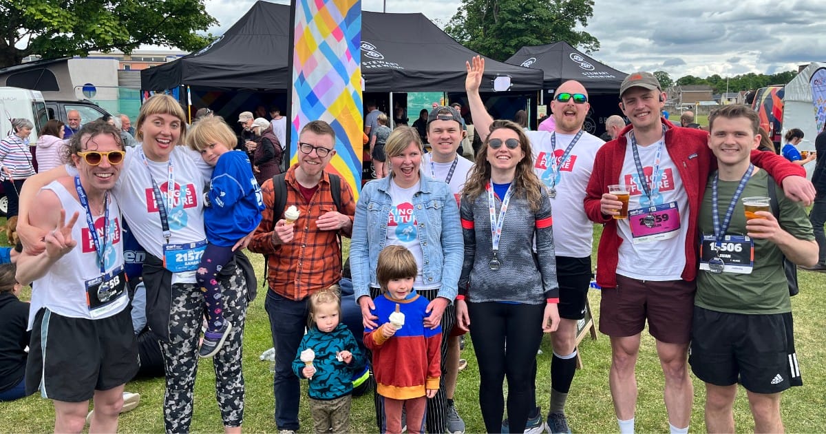 A group of people, some wearing medals and race bibs, pose outdoors at an event with tents and a crowd in the background. Two children stand at the front.