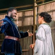 A man in a dark velvet coat gestures animatedly while speaking to a woman in a light dress on a wooden stage set.