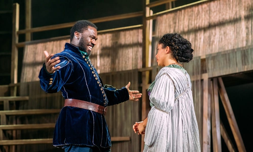 A man in a dark velvet coat gestures animatedly while speaking to a woman in a light dress on a wooden stage set.