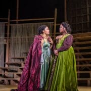 Two women in elaborate period costumes stand on a wooden stage set, engaged in conversation. One wears a red cloak, the other a green dress.